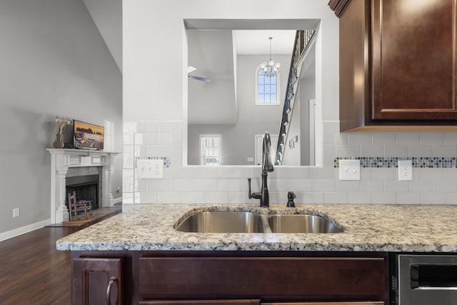 kitchen with a fireplace with raised hearth, light stone countertops, a sink, dark brown cabinets, and decorative backsplash