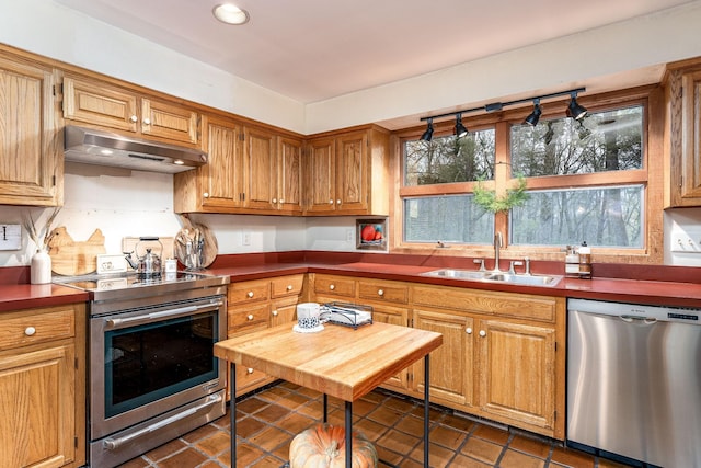 kitchen featuring dark countertops, appliances with stainless steel finishes, brown cabinetry, a sink, and under cabinet range hood