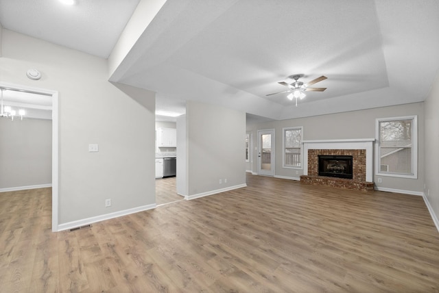 unfurnished living room featuring a tray ceiling, wood finished floors, a fireplace, and ceiling fan with notable chandelier