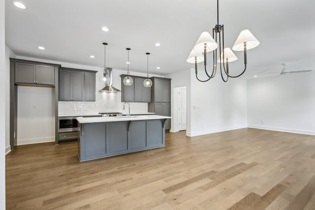 kitchen with hanging light fixtures, wall chimney range hood, a kitchen island with sink, and light countertops