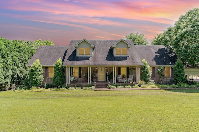 cape cod house with a porch, brick siding, a front lawn, and roof with shingles