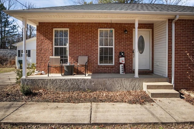 view of exterior entry with a porch, brick siding, and roof with shingles