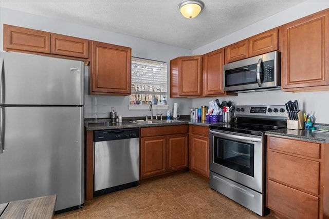 kitchen with brown cabinetry, dark stone counters, stainless steel appliances, a textured ceiling, and a sink