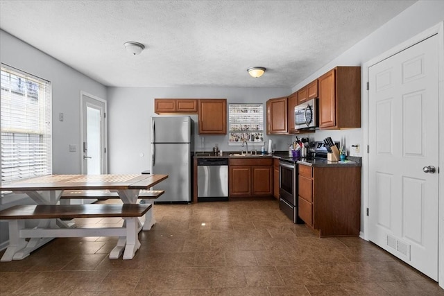 kitchen with brown cabinetry, dark countertops, appliances with stainless steel finishes, a textured ceiling, and a sink