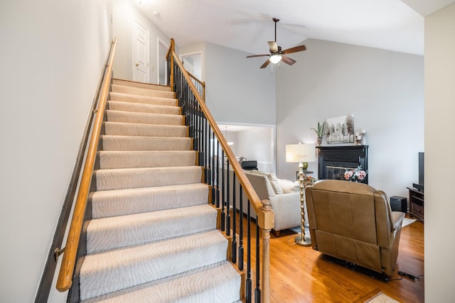 stairs with a ceiling fan, a glass covered fireplace, high vaulted ceiling, and wood finished floors