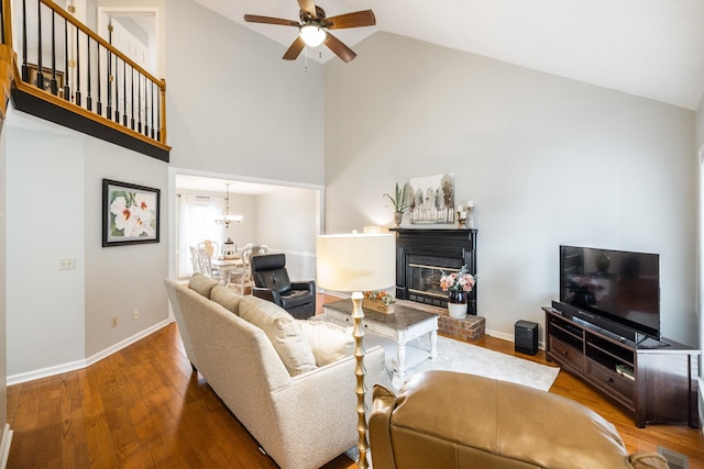 living area featuring a fireplace, wood finished floors, high vaulted ceiling, baseboards, and ceiling fan with notable chandelier