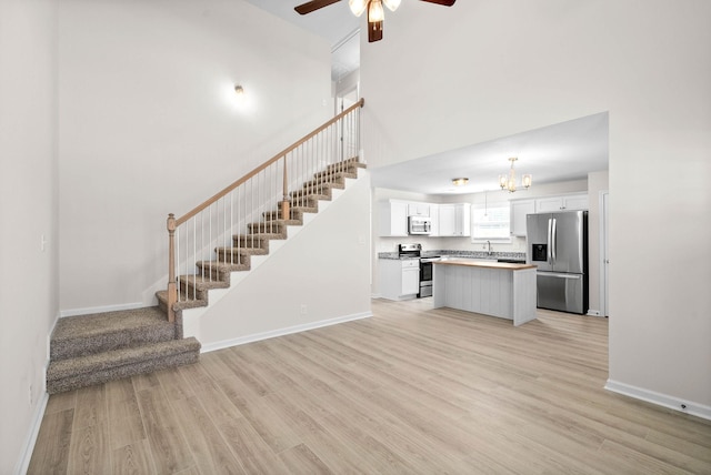 unfurnished living room featuring stairs, light wood finished floors, ceiling fan with notable chandelier, and baseboards