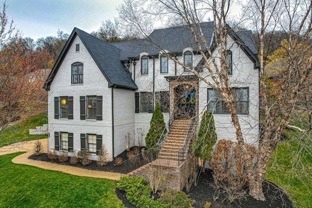 french country inspired facade with stairs, brick siding, roof with shingles, and a front yard