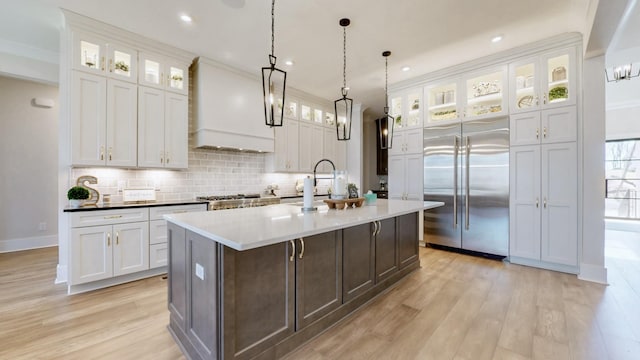 kitchen featuring an island with sink, custom range hood, a sink, stainless steel built in refrigerator, and backsplash