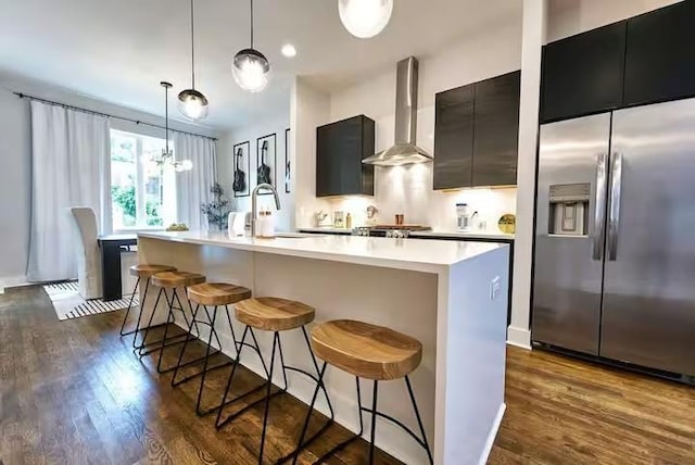 kitchen with an island with sink, wall chimney exhaust hood, a kitchen bar, pendant lighting, and built in fridge