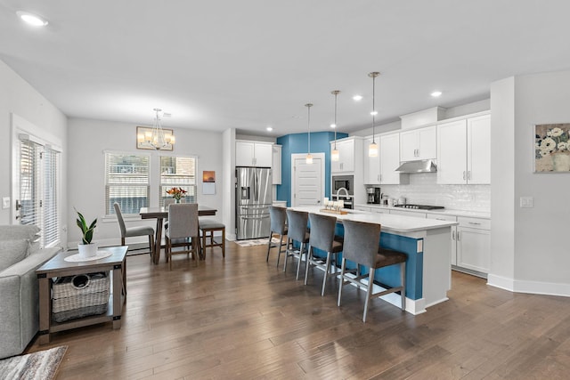 kitchen featuring pendant lighting, a center island with sink, stainless steel refrigerator with ice dispenser, light countertops, and under cabinet range hood