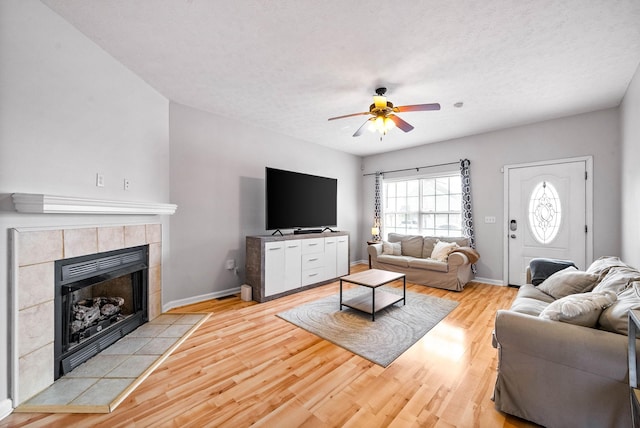 living area with a tiled fireplace, a ceiling fan, a textured ceiling, light wood-type flooring, and baseboards