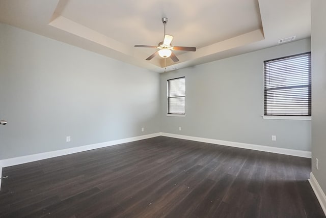 empty room featuring dark wood-type flooring, a ceiling fan, visible vents, baseboards, and a raised ceiling