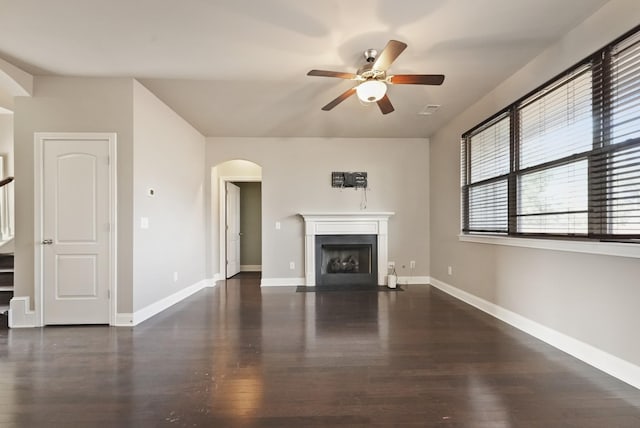 unfurnished living room featuring arched walkways, dark wood finished floors, a ceiling fan, baseboards, and a fireplace with flush hearth