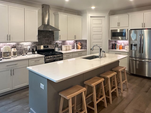 kitchen featuring a kitchen island with sink, a sink, light countertops, appliances with stainless steel finishes, and wall chimney range hood