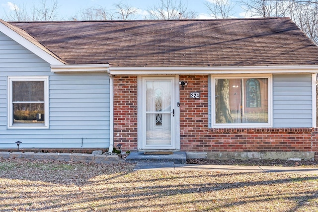 doorway to property featuring a shingled roof, brick siding, and a lawn