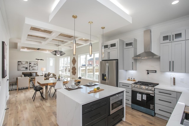 kitchen featuring coffered ceiling, wall chimney exhaust hood, appliances with stainless steel finishes, glass insert cabinets, and decorative light fixtures