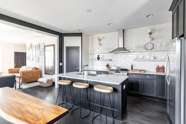 kitchen with open shelves, light countertops, appliances with stainless steel finishes, wall chimney range hood, and an island with sink