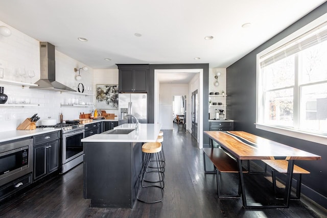 kitchen with an island with sink, stainless steel appliances, light countertops, wall chimney range hood, and open shelves