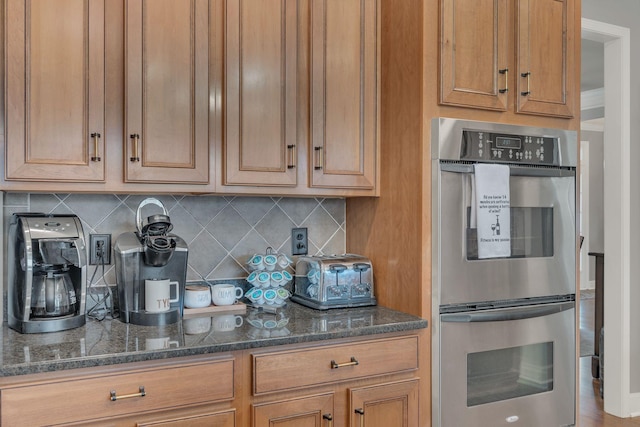 kitchen featuring tasteful backsplash, stainless steel double oven, brown cabinetry, and dark stone countertops