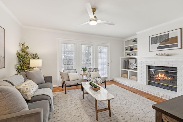 living area featuring built in features, crown molding, a ceiling fan, a brick fireplace, and light wood-type flooring