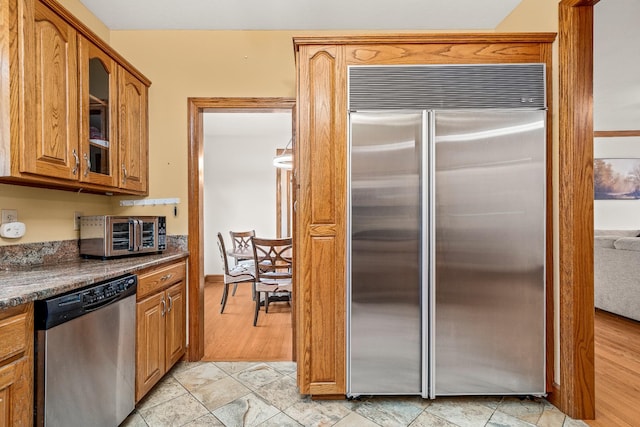 kitchen with a toaster, dark countertops, appliances with stainless steel finishes, brown cabinetry, and glass insert cabinets
