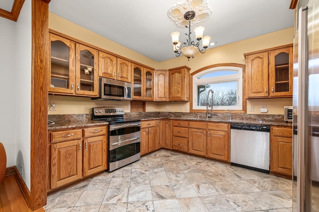 kitchen with stainless steel appliances, a sink, brown cabinetry, glass insert cabinets, and an inviting chandelier
