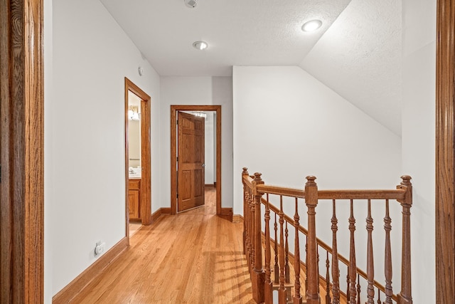 corridor with a textured ceiling, an upstairs landing, baseboards, vaulted ceiling, and light wood finished floors