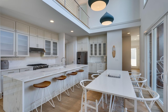 kitchen with a kitchen island with sink, under cabinet range hood, stainless steel appliances, a sink, and a kitchen bar