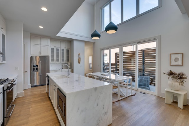 kitchen with appliances with stainless steel finishes, glass insert cabinets, a sink, an island with sink, and light wood-type flooring