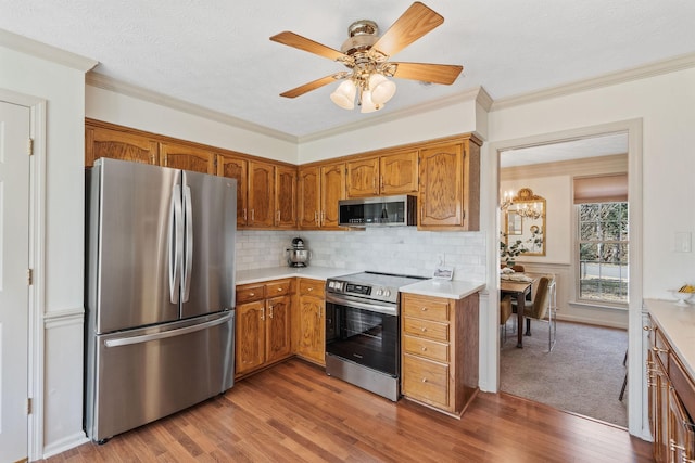 kitchen with stainless steel appliances, wood finished floors, light countertops, brown cabinetry, and crown molding