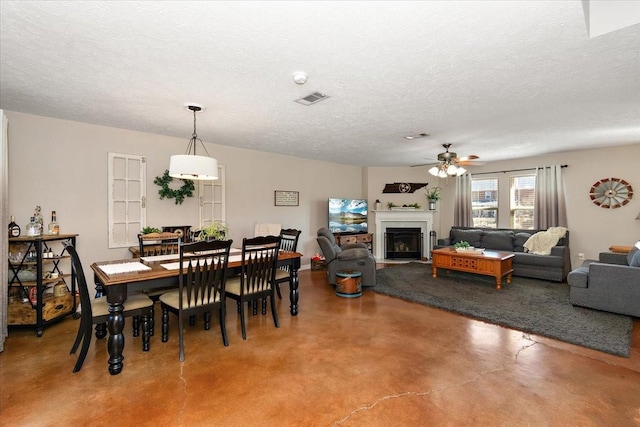 dining area featuring finished concrete flooring, visible vents, a fireplace with flush hearth, a ceiling fan, and a textured ceiling