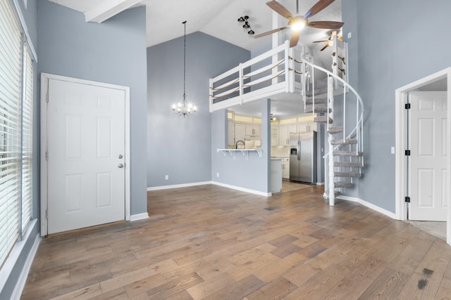 unfurnished living room featuring high vaulted ceiling, ceiling fan with notable chandelier, wood finished floors, baseboards, and stairs