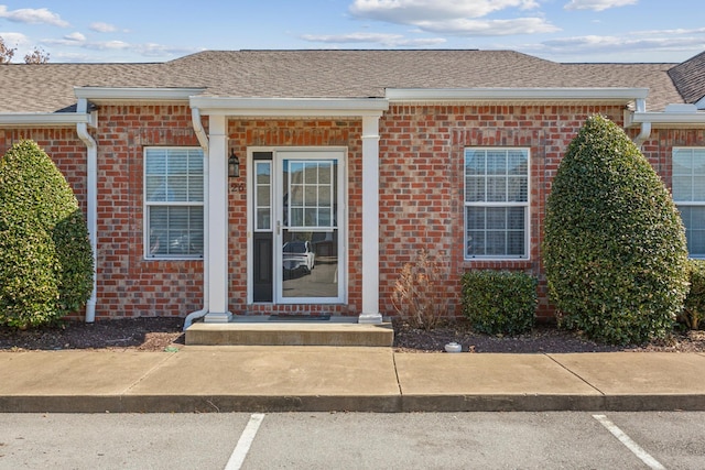 doorway to property with uncovered parking, brick siding, and roof with shingles