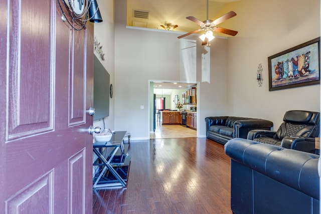 living area featuring wood finished floors, a towering ceiling, a ceiling fan, visible vents, and baseboards