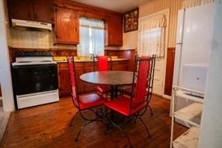 kitchen with electric range oven, brown cabinets, wood finished floors, freestanding refrigerator, and under cabinet range hood