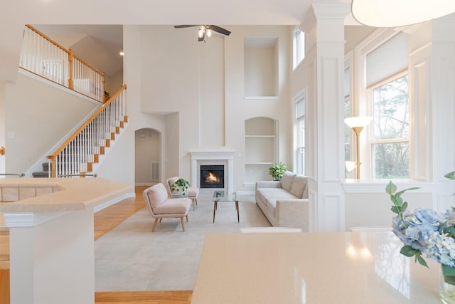 living room with arched walkways, a ceiling fan, a lit fireplace, a high ceiling, and built in shelves