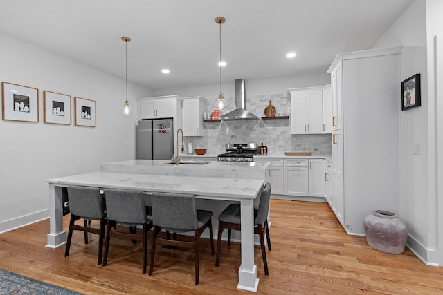 kitchen with light wood finished floors, an island with sink, appliances with stainless steel finishes, wall chimney range hood, and a sink