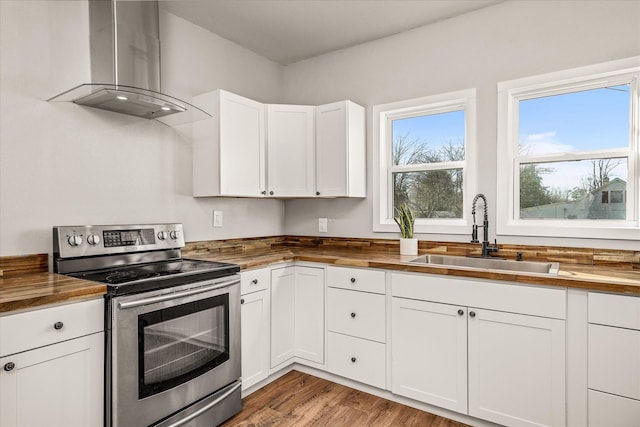 kitchen with white cabinets, wall chimney range hood, stainless steel range with electric stovetop, wooden counters, and a sink