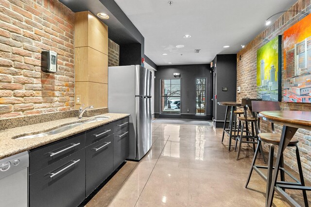 kitchen with stainless steel appliances, modern cabinets, a sink, brick wall, and light stone countertops