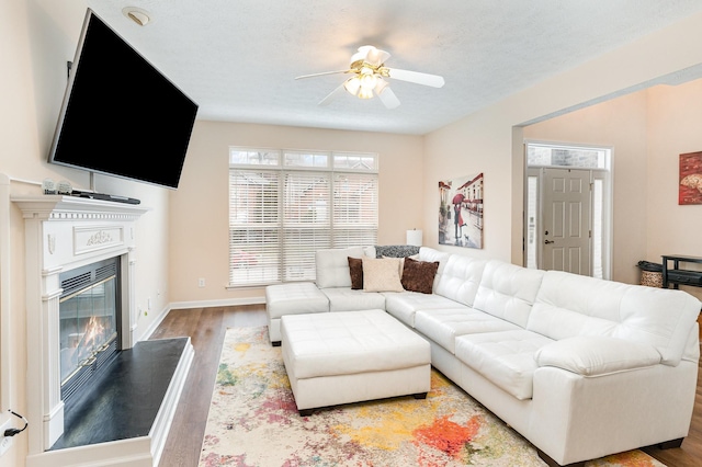 living room featuring baseboards, a ceiling fan, a glass covered fireplace, wood finished floors, and a textured ceiling