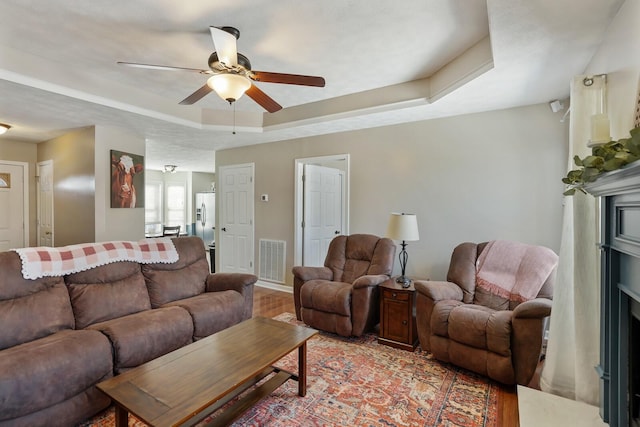 living room with a fireplace, a raised ceiling, visible vents, light wood-style floors, and ceiling fan