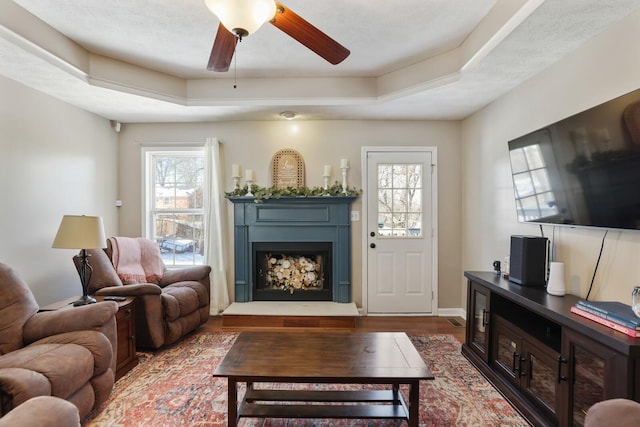 living room with a wealth of natural light, dark wood-style flooring, a fireplace with raised hearth, and a tray ceiling