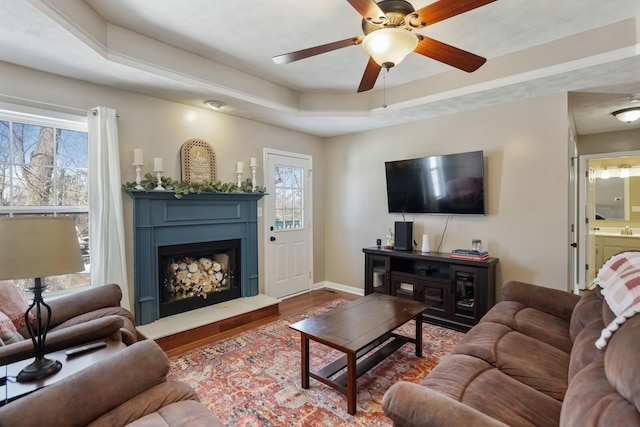 living area with a tray ceiling, a fireplace with raised hearth, a ceiling fan, wood finished floors, and baseboards