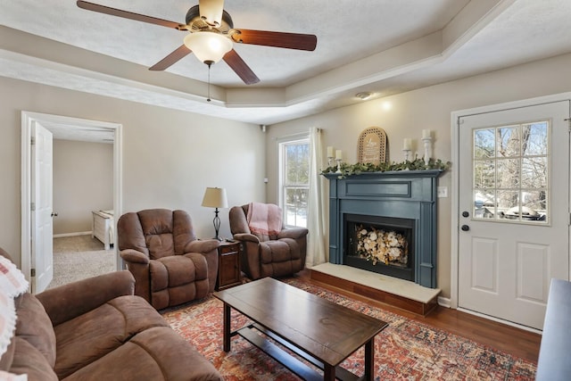 living room featuring a tray ceiling, a fireplace with raised hearth, a ceiling fan, wood finished floors, and baseboards