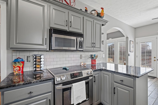 kitchen with gray cabinetry, a peninsula, appliances with stainless steel finishes, decorative backsplash, and dark stone counters