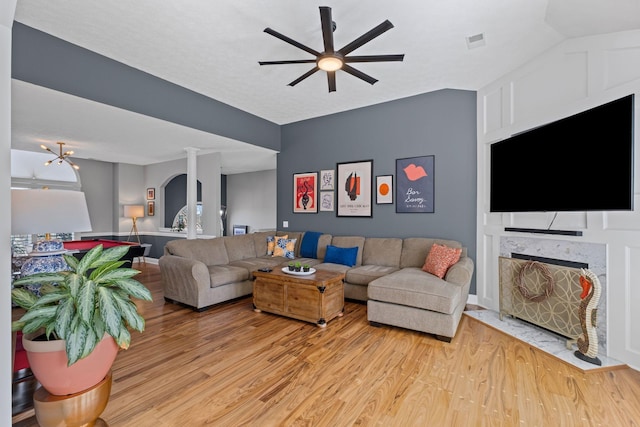 living room with ceiling fan, a fireplace with flush hearth, visible vents, light wood-style floors, and ornate columns