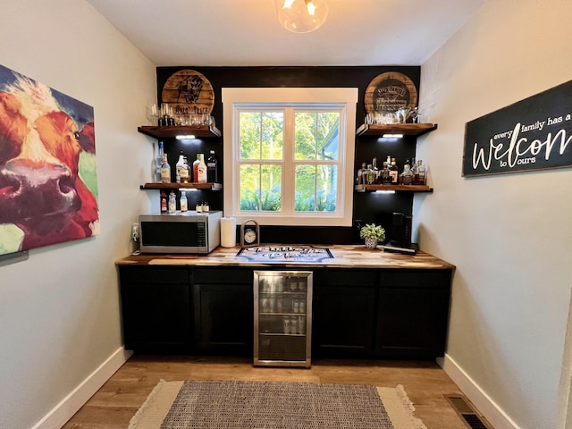 bar with baseboards, light wood-type flooring, wine cooler, stainless steel microwave, and a bar