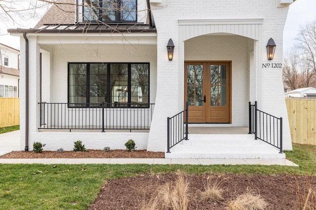 doorway to property featuring french doors, brick siding, fence, and covered porch