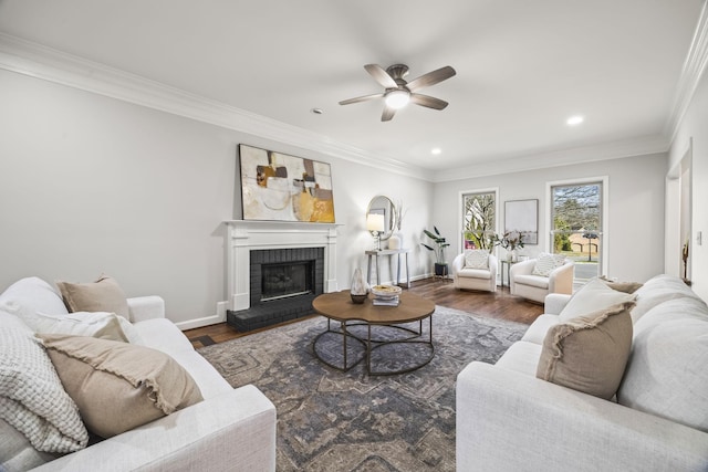 living room featuring a ceiling fan, wood finished floors, crown molding, baseboards, and a brick fireplace